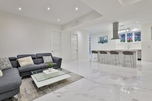 A modern living room with a grey sofa, a glass coffee table, and a kitchen area with a bar and stools.