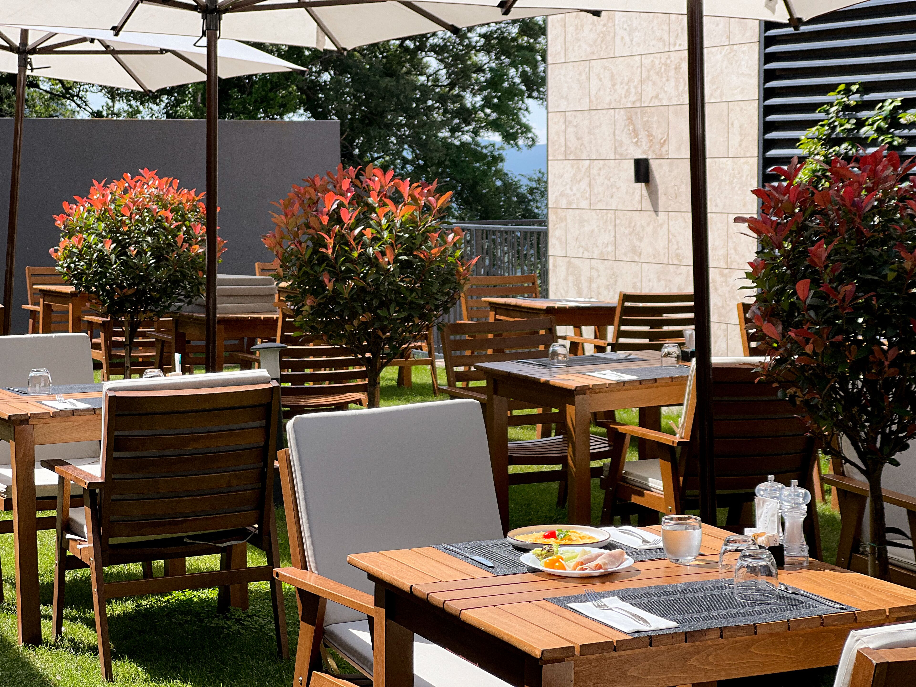 An outdoor dining area with wooden tables and chairs, surrounded by potted plants and umbrellas.