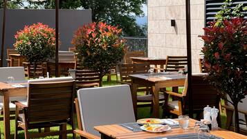 Un área de comedor al aire libre con mesas y sillas de madera, rodeada de plantas en macetas y sombrillas.