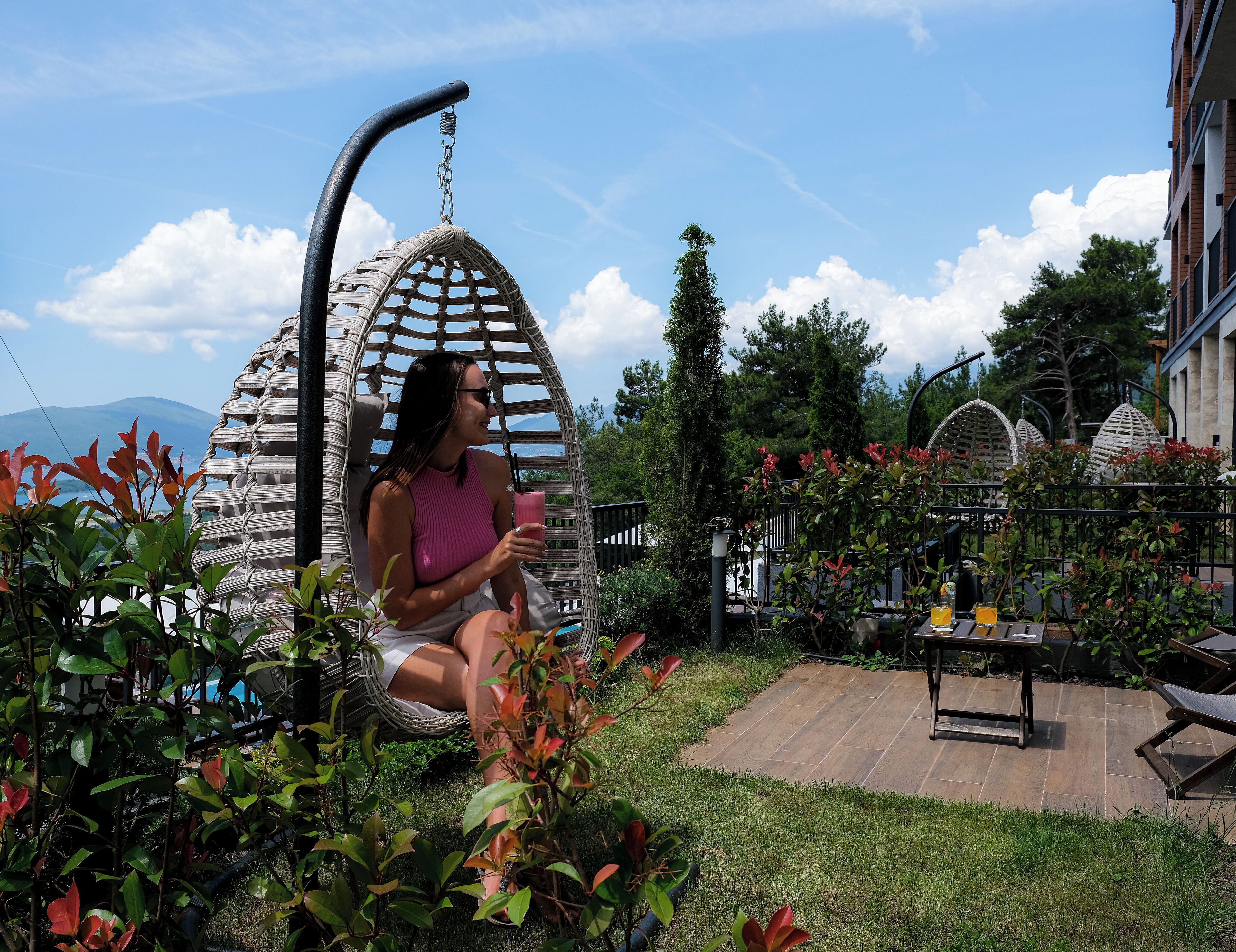 A person sitting in a hanging chair in a garden area with a table and chairs nearby.