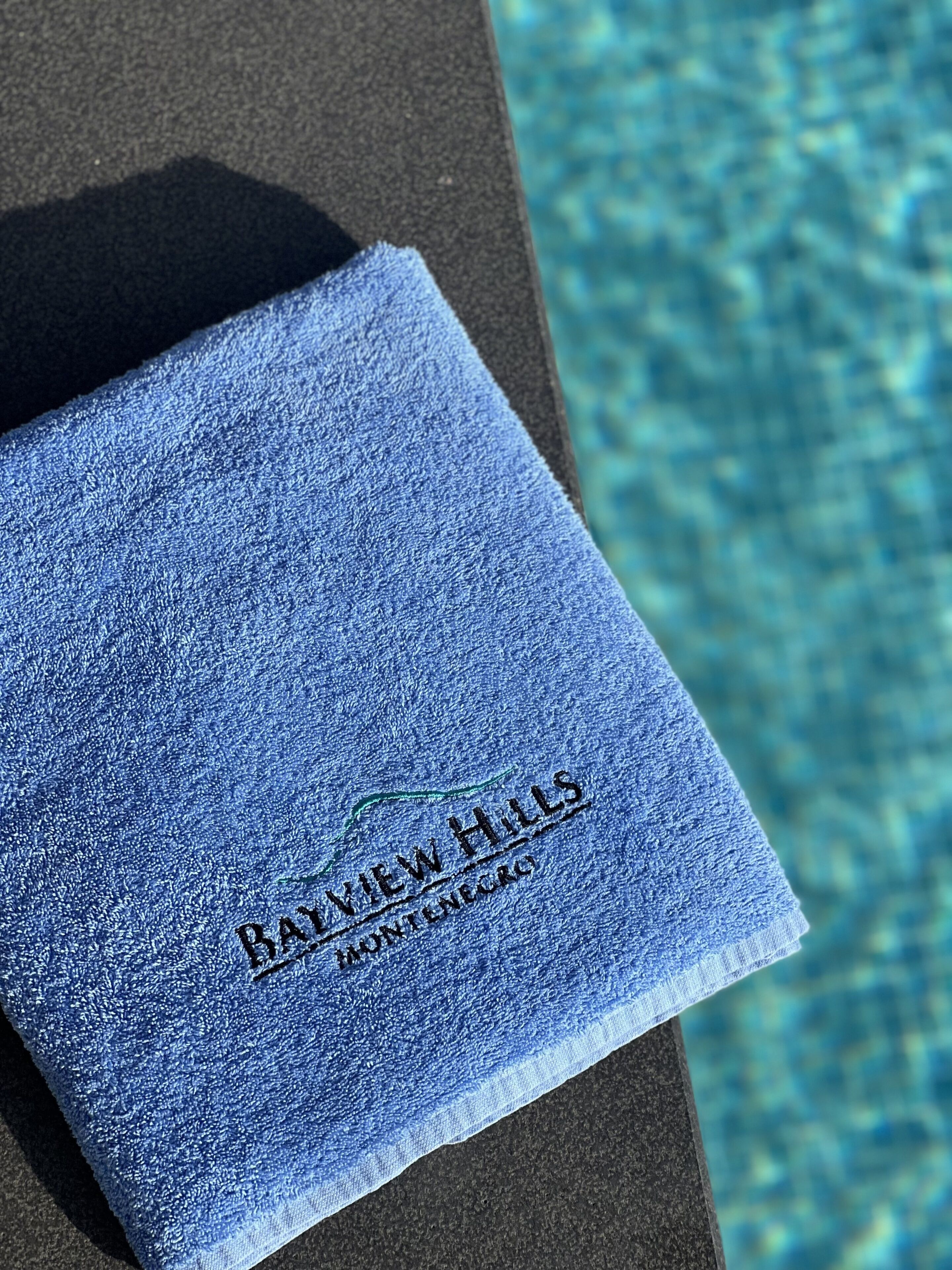 A blue towel with "RAYVIEW HILLS" and "Montgomery" embroidered on it, placed on a dark surface near a pool.