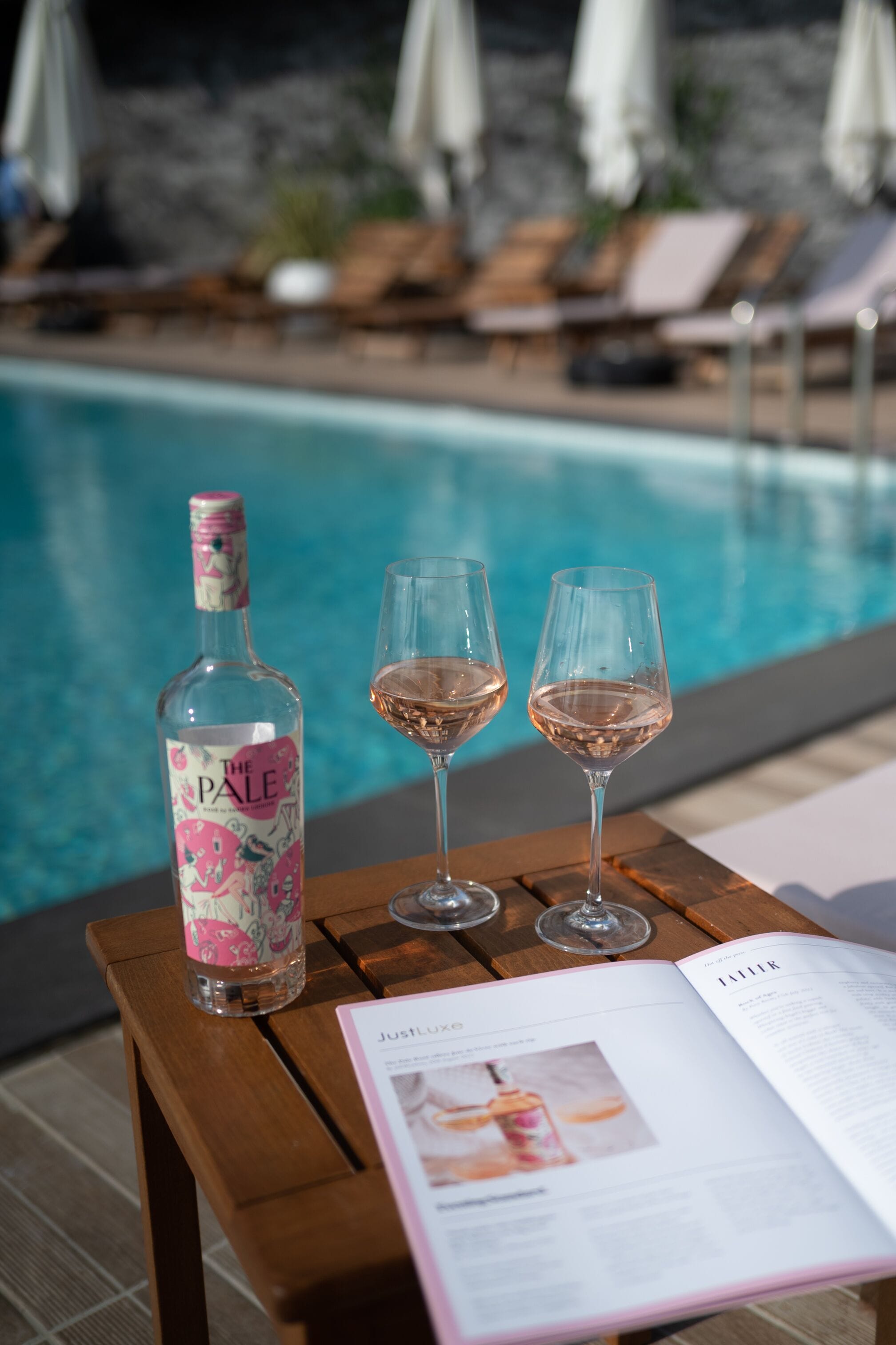 Una bottiglia di The Pale, due bicchieri di vino e una rivista su un tavolo di legno vicino a una piscina.