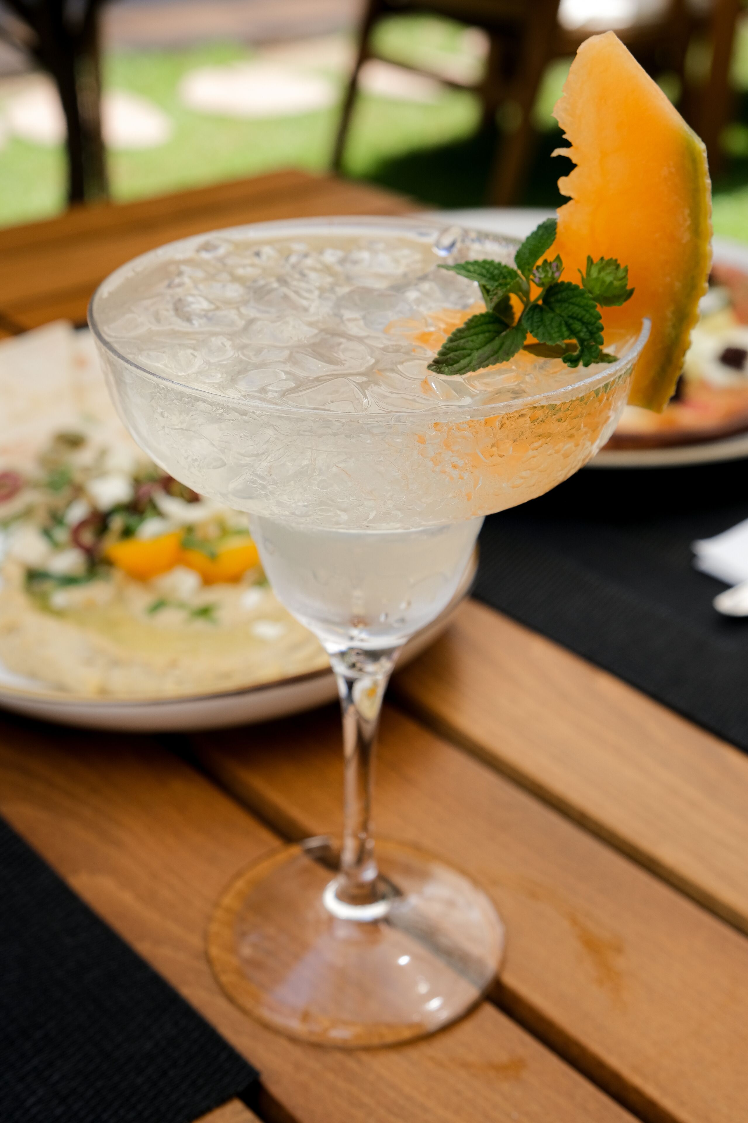 Un verre transparent contenant une boisson fraîche avec des glaçons, agrémenté d’une tranche de fruit et d’une feuille de menthe, posé sur une table en bois.