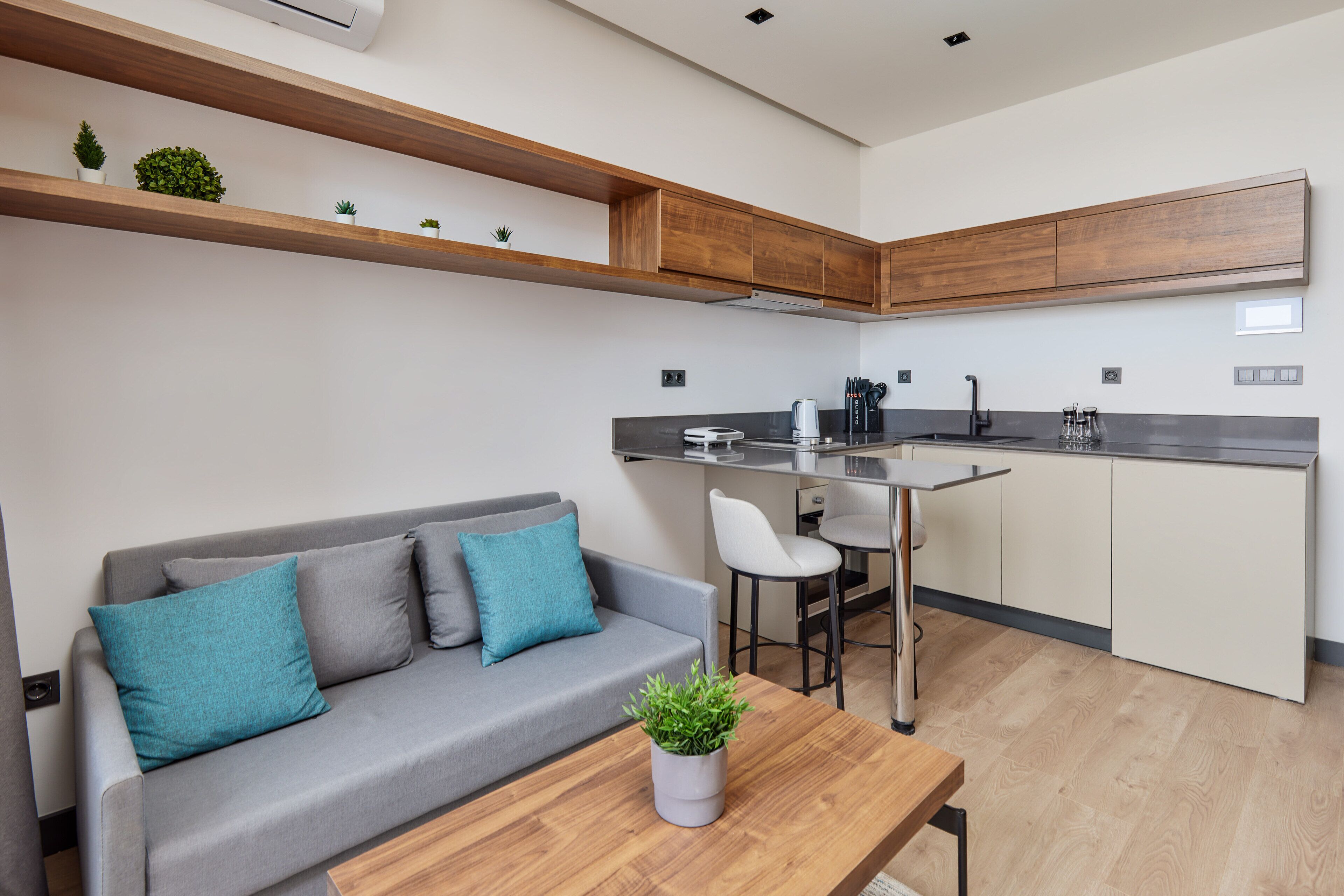 A modern kitchen with wooden cabinets, a grey sofa with teal cushions, and a small table with a plant.