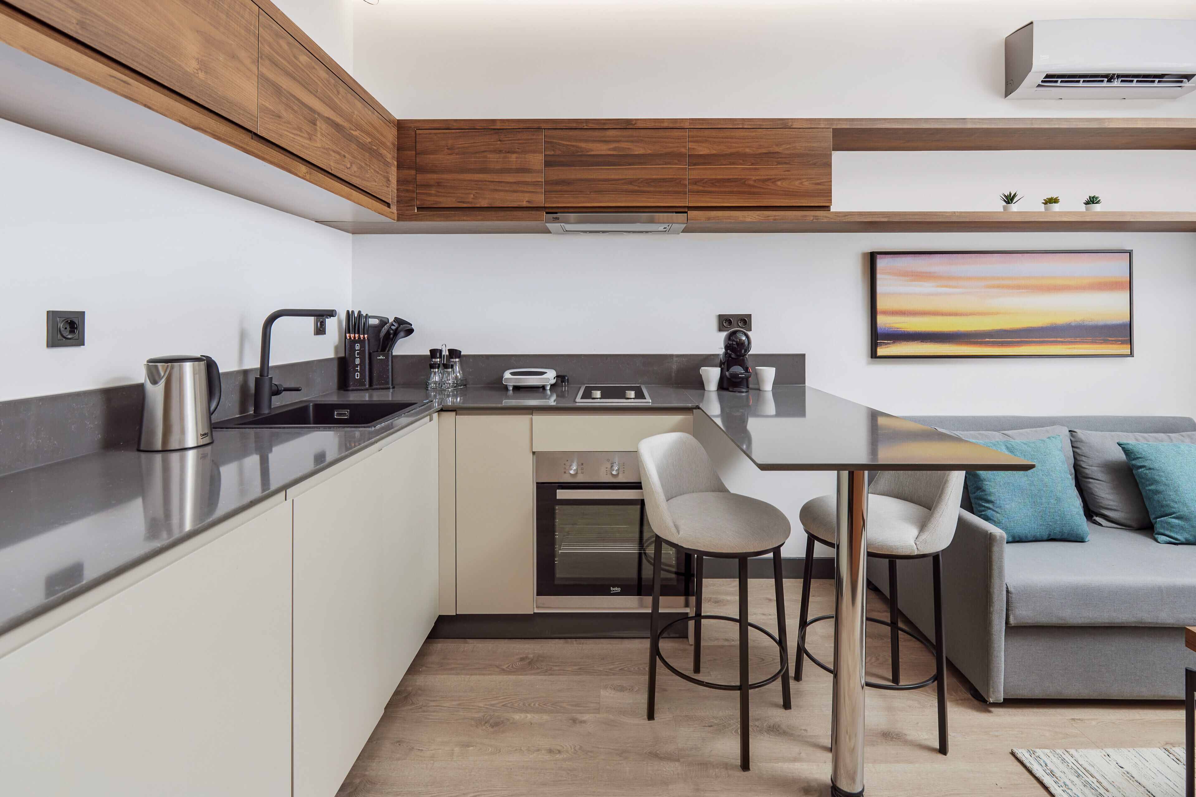 A modern kitchen with a built-in oven, a coffee machine, and a dining area with a grey sofa and two bar stools.