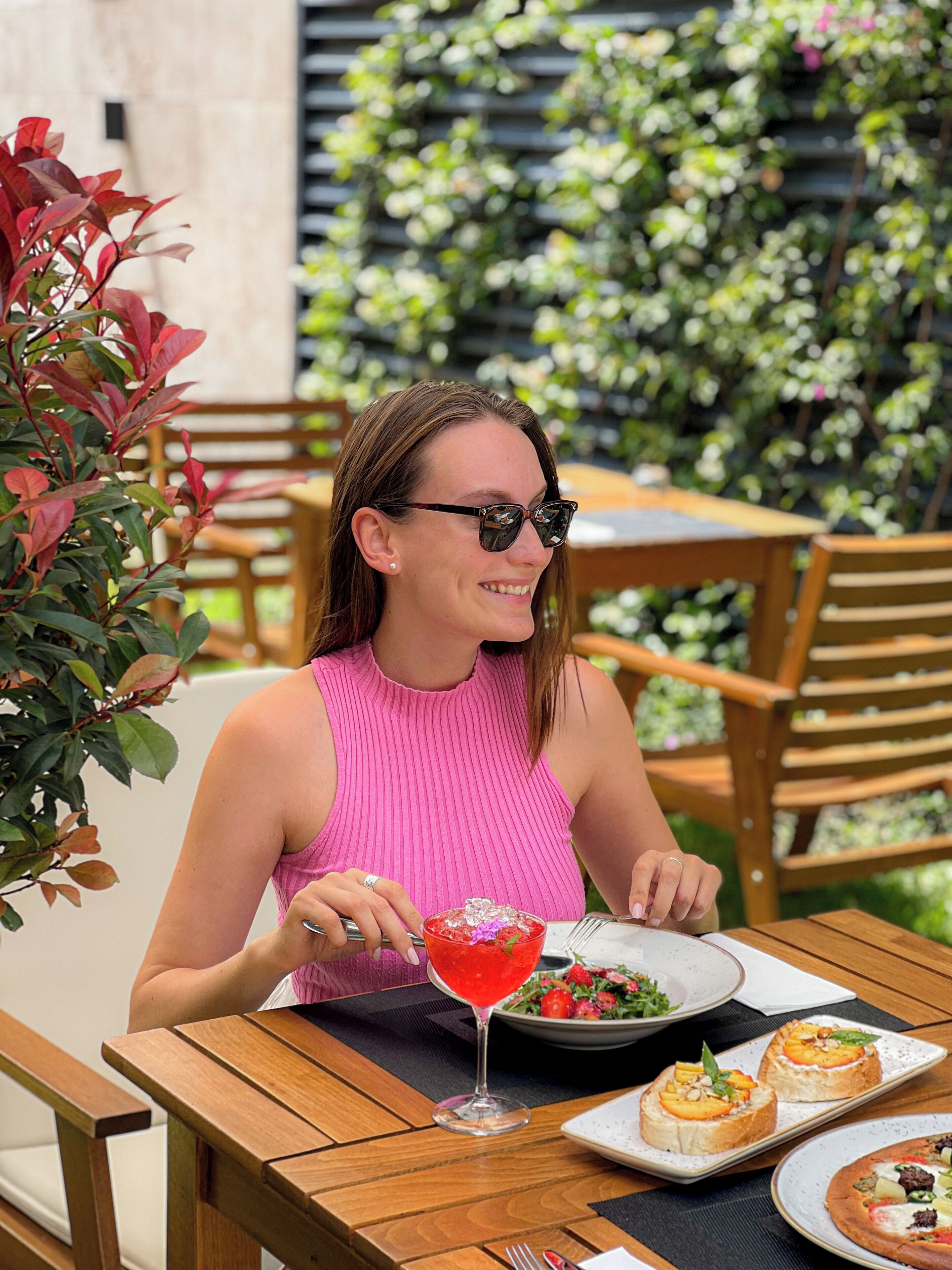 A person seated at an outdoor table with a drink, a plate of food, and a dessert plate.