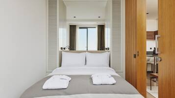 Una habitación de hotel con una cama grande, dos toallas blancas, un plaid gris y puertas de madera que dan a una kitchenette.