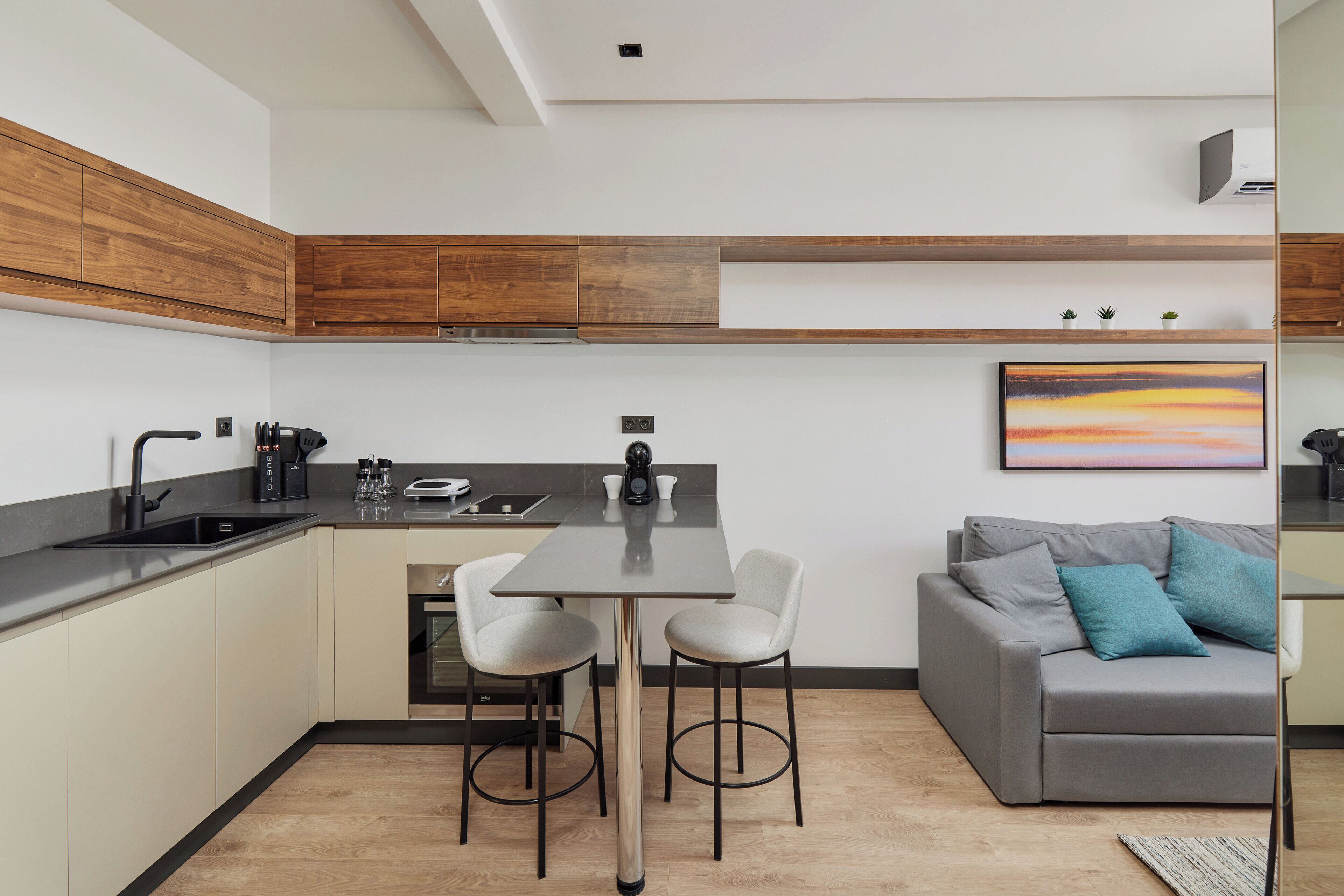 A modern kitchen with a dining area, wooden cabinets, a grey sofa with blue cushions, and a painting on the wall.