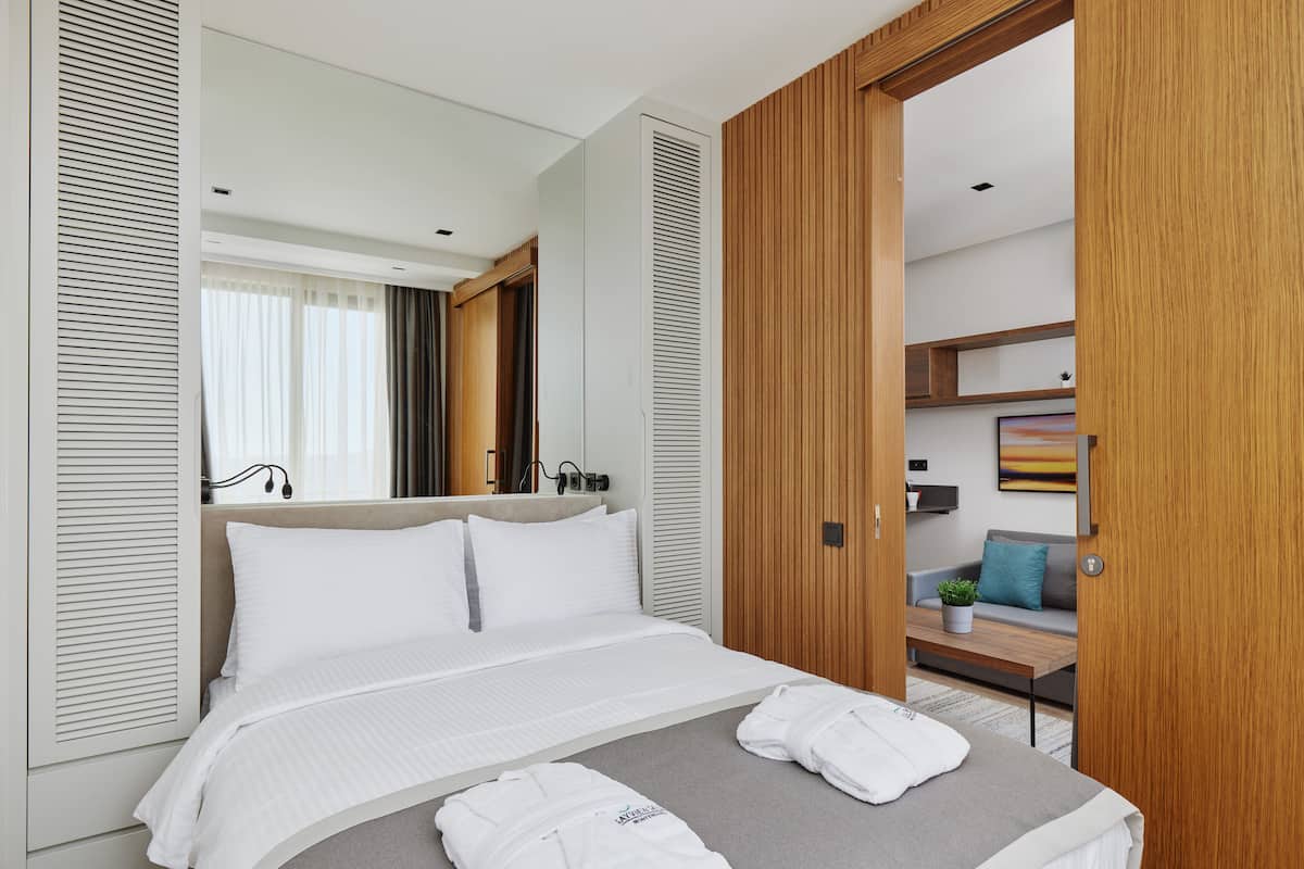 A modern hotel room with a large bed, a wooden wardrobe, a small seating area, and a view into another room.