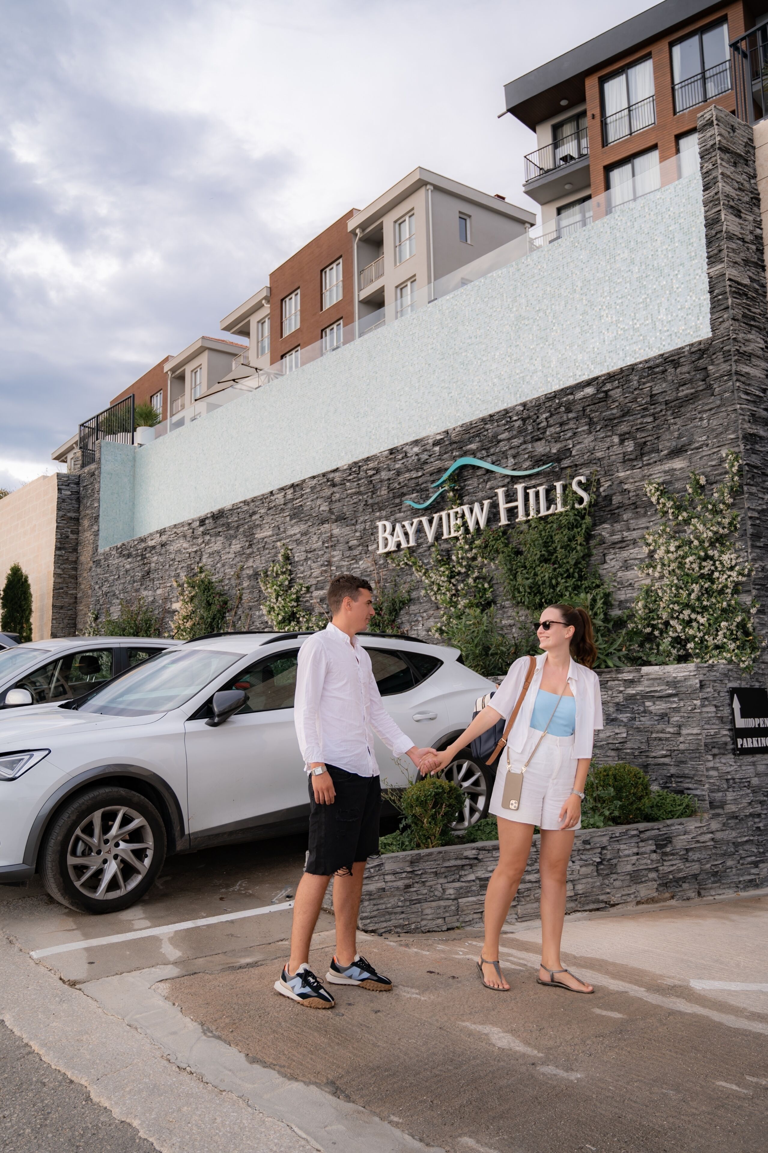 Una pareja tomándose de las manos frente a un edificio con fachada de piedra y un letrero que dice 'Bayview Hills'.