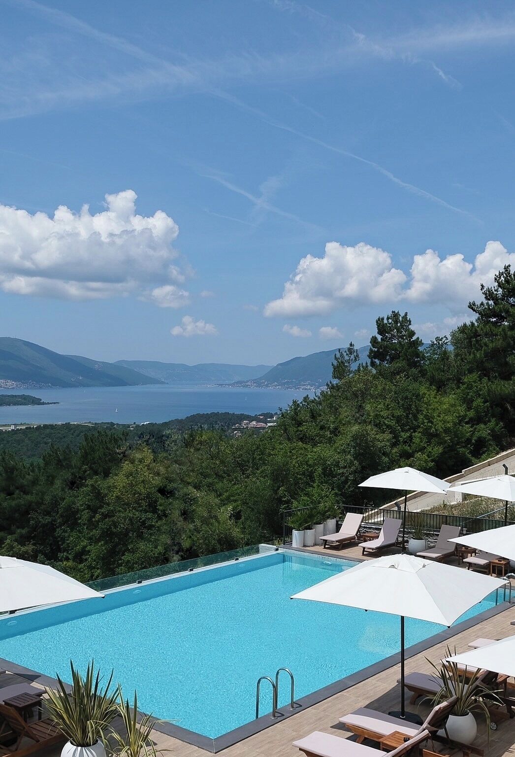Piscina con sillas de descanso y sombrillas, rodeada de exuberante vegetación y vistas a un lago y montañas.
