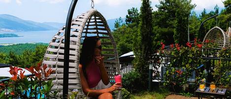 A person sitting in a hanging chair in a garden with a view of a lake and mountains.