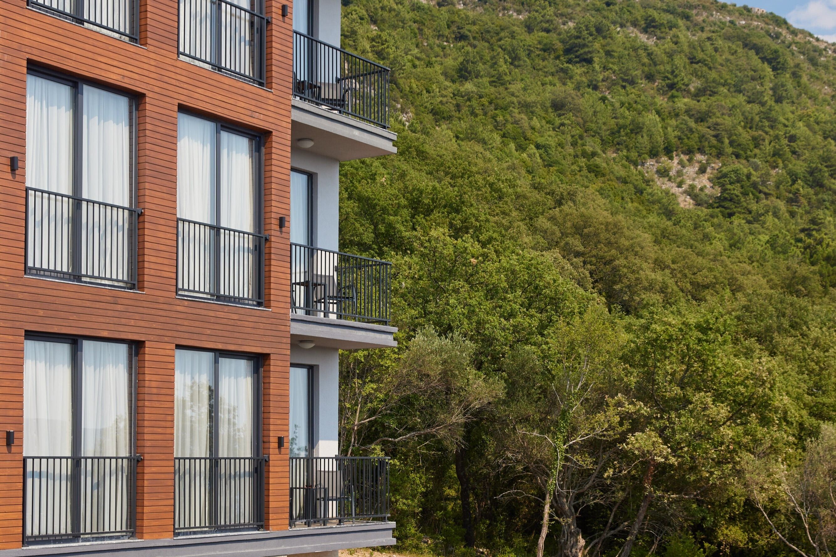 A multi-story building with balconies, large windows, and a forested hillside in the background.
