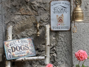 Una pared rústica con tres letreros: uno que dice “UN HOGAR NO ES UN SOGAR SIN UN PERRO” con un Yorkshire Terrier, otro que reza “SOLO UNA CHICA A LA QUE LE ENCANAN LOS PERROS Y LA JARDINERÍA” con un perro y flores, y un tercero con una campana y un perro.