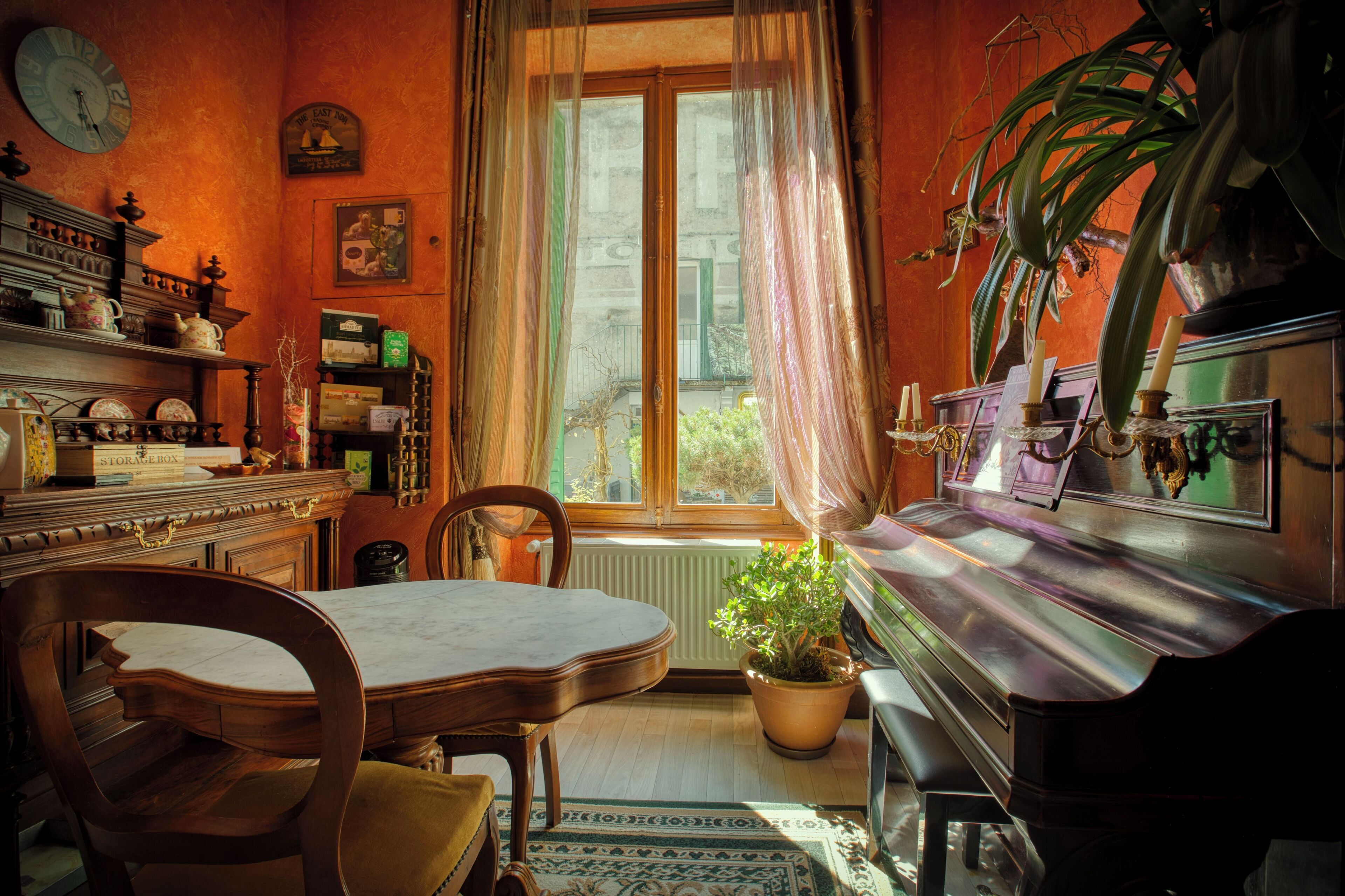 Een kamer met een houten tafel, stoelen, een radiator, een raam met gordijnen, een kamerplant en een klok aan de muur.