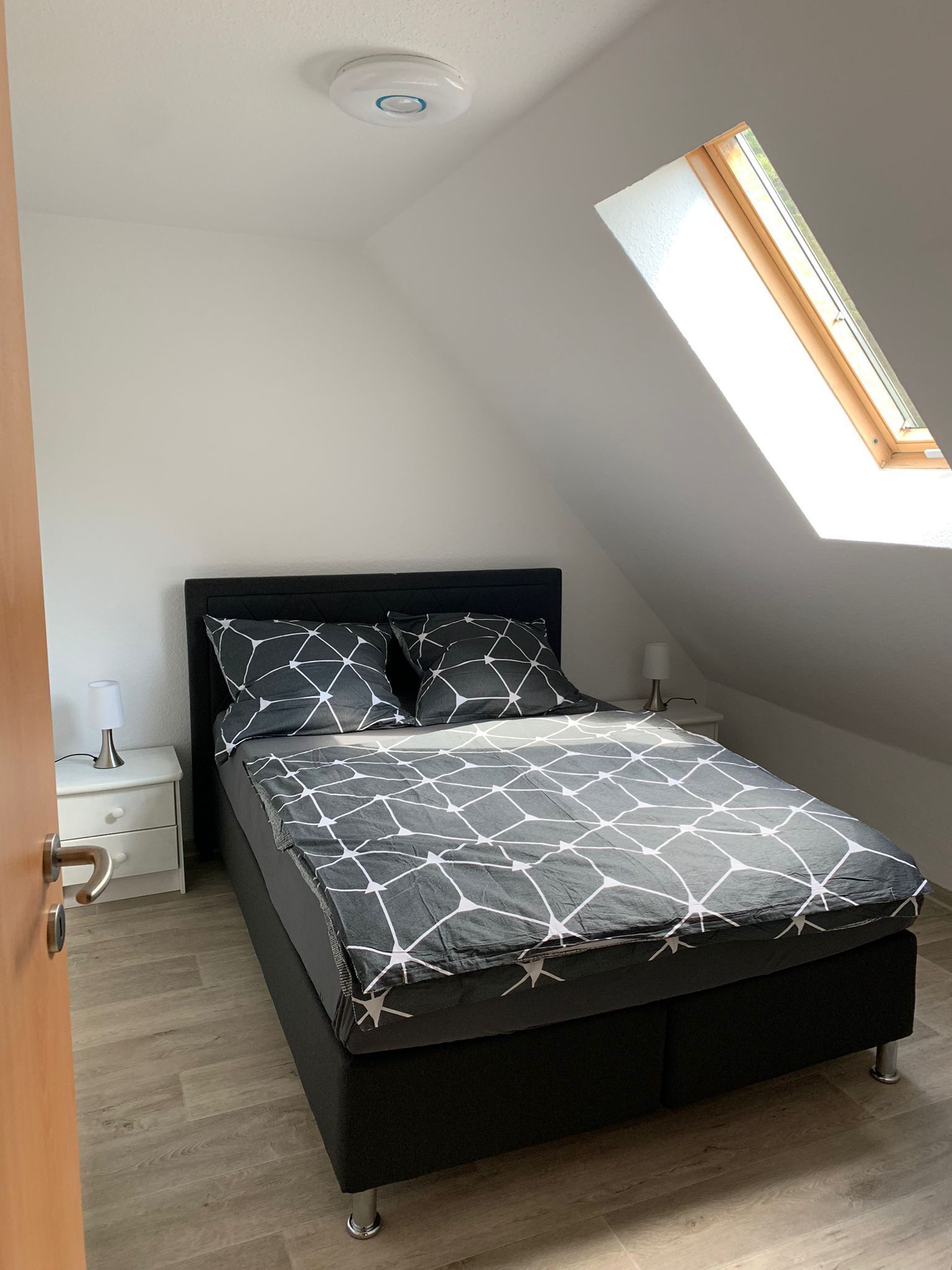 A bedroom with a sloped ceiling, a skylight, a bed with a geometric-patterned bedspread, a bedside table with a lamp, and a nightstand.