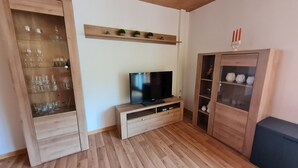 Een woonkamer met een houten vloer, een tv op een houten standaard, een glazen kast met wijnglazen, en een kast met decoratieve objecten.