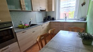 Een keuken met houten keukenkastjes, een eettafel, stoelen, een fornuis en een raam met uitzicht op gebouwen.