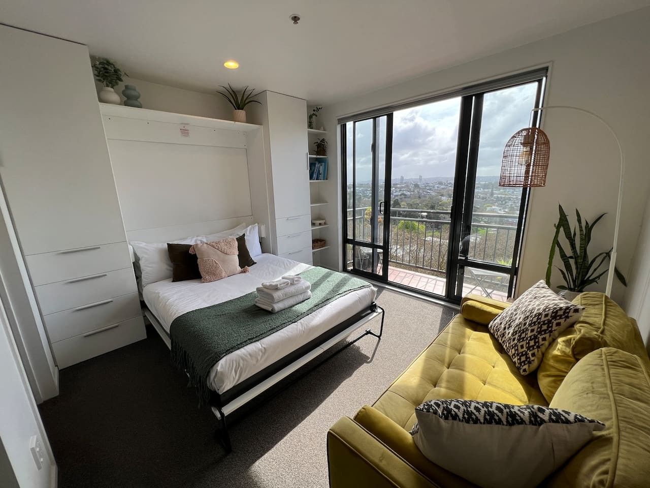A modern bedroom with a bed, a yellow sofa, and a large window with a city view.