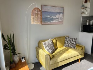 A yellow sofa with patterned cushions, a woven floor lamp, a potted plant, and a framed picture of a beach scene. - Cosy studio with balcony and amazing views! (Auckland)