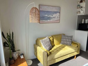 A yellow sofa with patterned cushions, a floor lamp, a potted plant, and a painting of a beach scene.