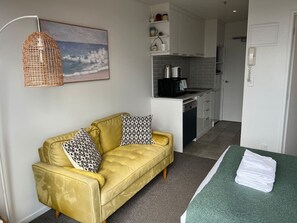 A yellow sofa with patterned cushions, a green towel on a table, and a kitchenette in the background. - Cosy studio with balcony and amazing views! (Auckland)