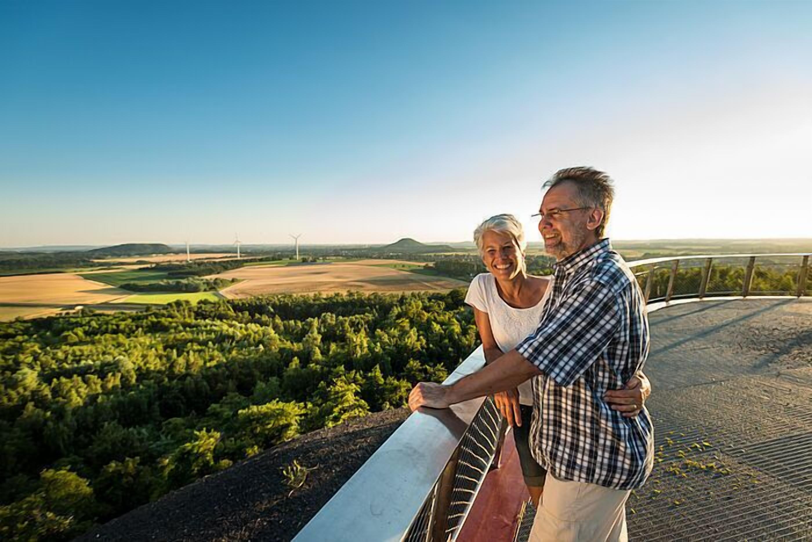 A couple standing on a balcony overlooking a scenic landscape with wind turbines in the distance.