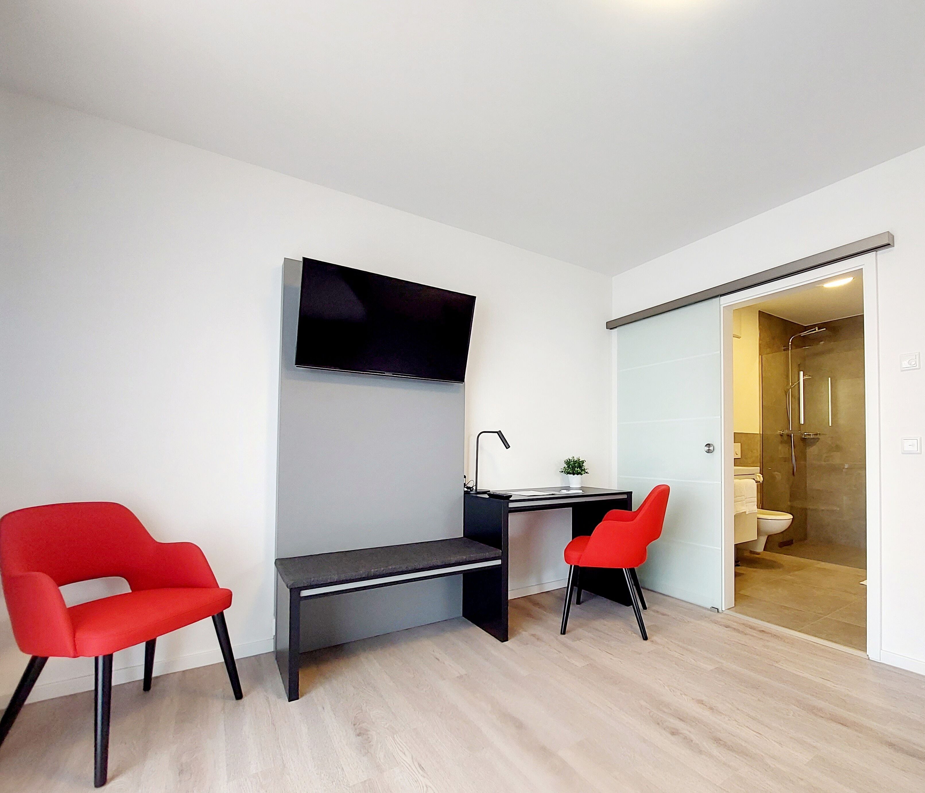 Una habitación moderna con escritorio, dos sillas rojas, televisor y puerta corredera al baño.