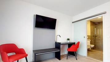 Una habitación moderna con escritorio, dos sillas rojas, televisor y puerta corredera al baño.
