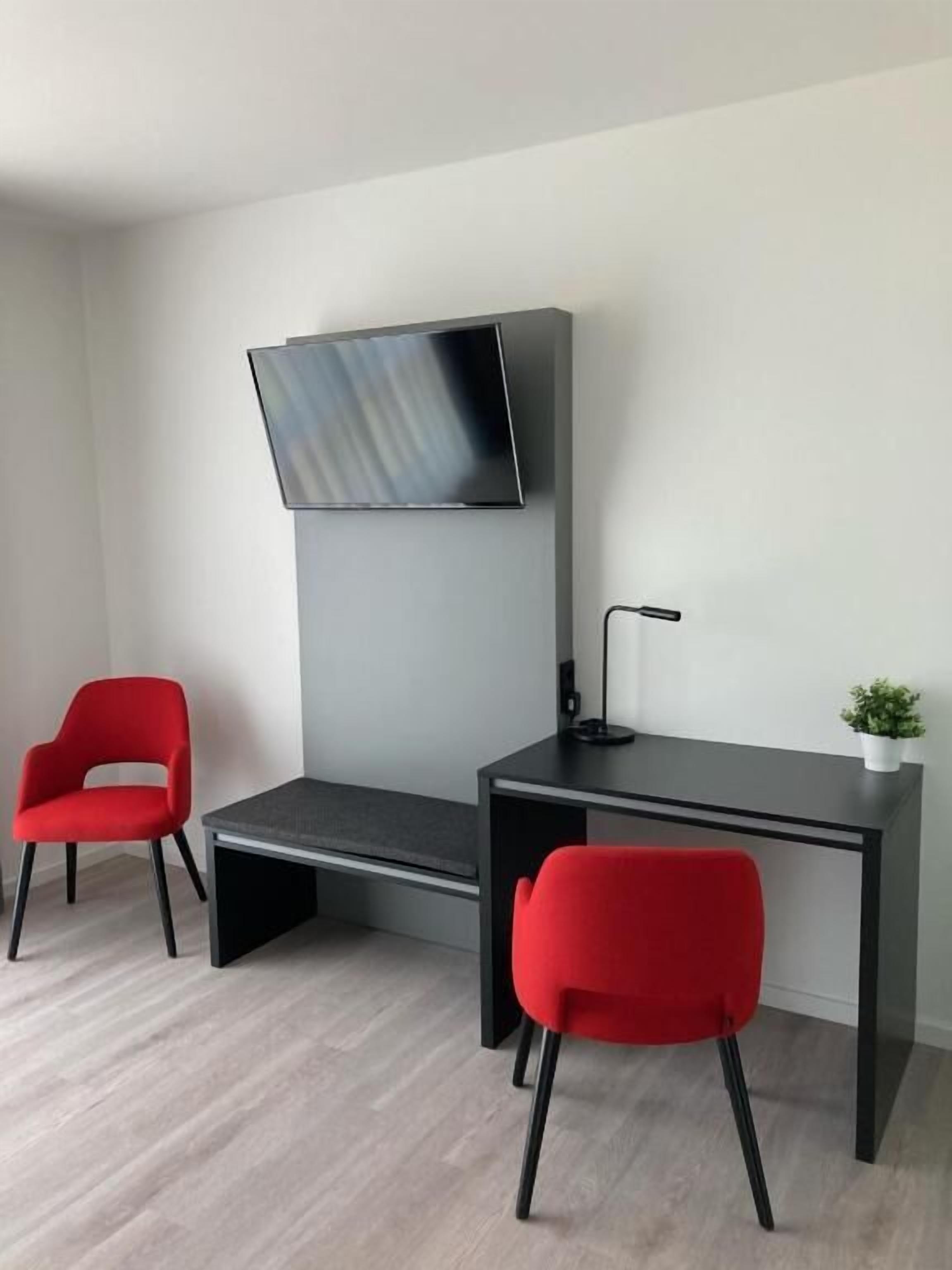 Ein modernes Zimmer mit Schreibtisch, zwei roten Stühlen, einem an der Wand befestigten Fernseher und einer kleinen Pflanze auf dem Schreibtisch.