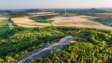Vista aérea de una zona rural con un camino sinuoso, un bosque denso y aerogeneradores a lo lejos.