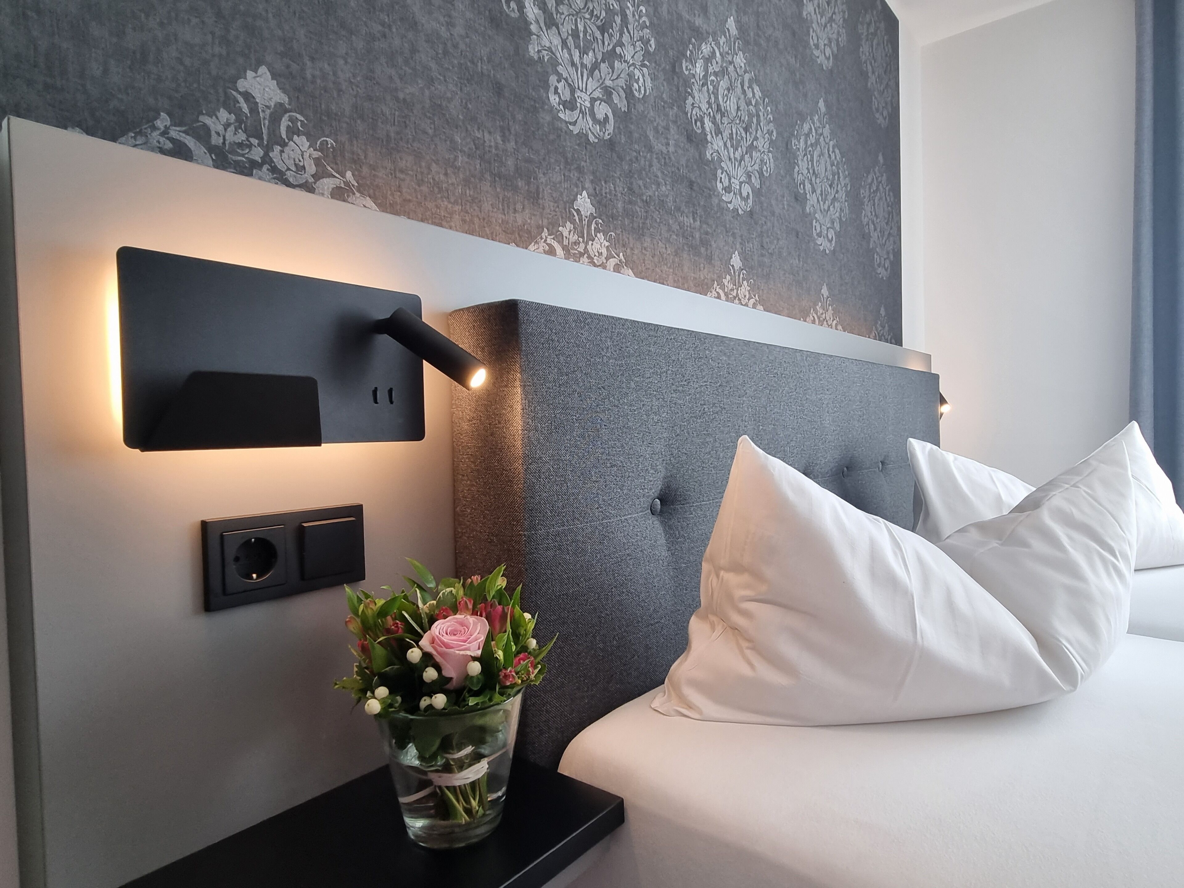 Una habitación de hotel moderna con un cabecero gris con botonadura, ropa de cama blanca, una mesita de noche negra con una lámpara de pared, y un jarrón con flores.