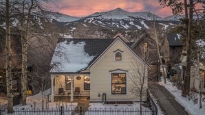 Una casa de dos pisos con techo cubierto de nieve, un porche con sillas y montañas al fondo.