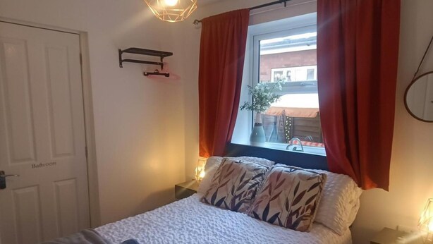 A bedroom with a bed, a window with red curtains, a bedside table with a lamp, and a wall-mounted shelf. - Beach Apartment With Garden (Skegness)