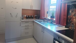 A compact kitchen with white cabinets, a stainless steel sink, and a window with pink curtains. - Beach Apartment With Garden (Skegness)