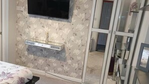 A bedroom with a TV mounted on the wall, floral wallpaper, a bedside table, and a glass door leading to another room. - Beach Apartment With Garden (Skegness)