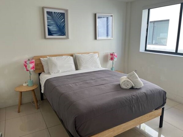 A neatly made bed with a grey duvet, two white pillows, and a rolled towel on the footboard. There are two bedside tables with lamps, and the room has a large window with a view of the sky. - Miami Suite 201 (Miami)