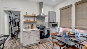 A modern kitchen with white cabinetry, a built-in oven, microwave, and a stainless steel range hood. There is a dining area with a table set for two, and a window with blinds. - Contemporary Apartment - 5 Min To TIAA Bank Field! (Jacksonville)