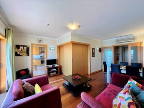 A modern living room with a wooden floor, a TV on a stand, and a dining area with a table and chairs.