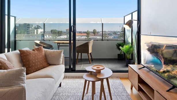 Ein modernes Wohnzimmer mit einer Couch, einem kleinen Tisch und einem Fernseher. Es gibt einen Balkon mit Blick auf die Stadt und eine Pflanze.