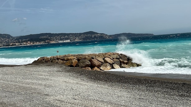 A rocky shoreline with waves crashing against it, a clear sky, and a distant view of a coastal town.