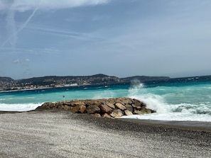 A rocky shoreline with waves crashing against it, a clear sky, and a distant view of a coastal town. - Swimming pool apartment near the sea French Riviera:  Cannes, Nice and Monaco (Cagnes-sur-Mer)