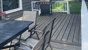 A deck with a grill, outdoor seating, and a house with a large window and a door. - Entire Home with Lakeview (L'Anse)