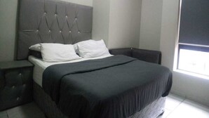 A bedroom with a tufted headboard, a bed with a black bedspread, white pillows, and a nightstand. - Central Rosebank Unit (Johannesburg)
