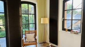A room with a wooden chair, a lamp, a small table with a book and a remote control, and large windows with a view of greenery and buildings. - Charming rear building near the old town (Osnabrück)
