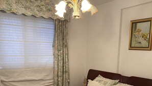 A bedroom with a floral-patterned bedspread, a wooden headboard, a chandelier, a painting on the wall, and a window with curtains. - Apartamento Playa Ladeira (Santa Cristina da Ramallosa)