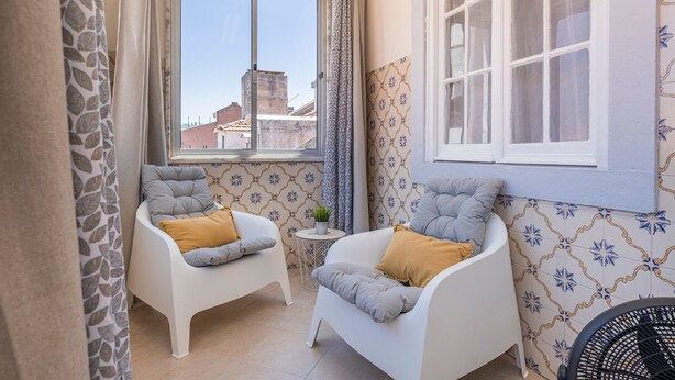 Una habitació petita i acollidora amb dues butaques blanques i una taula rodona, una finestra amb cortines i rajoles de parets amb dibuixos.