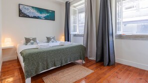 Un lit bien fait, recouvert d’une couverture verte, un tableau représentant une vague accroché au mur et une fenêtre avec des rideaux gris.