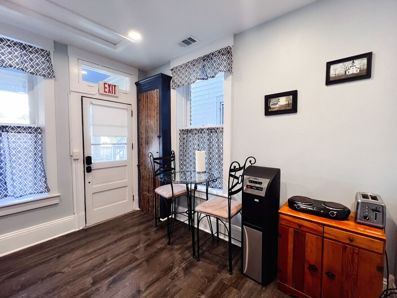 Quarto com piso de madeira, porta branca com a placa 'EXIT', pequena mesa com duas cadeiras, armário com rádio e torradeira, e duas fotos emolduradas na parede.