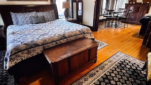 Ein Schlafzimmer mit einem Bett, einer hölzernen Bank, einem Teppich, einem Gemälde, einem Spiegel, einer Kommode und einem Essbereich mit Stühlen und einem Tisch.